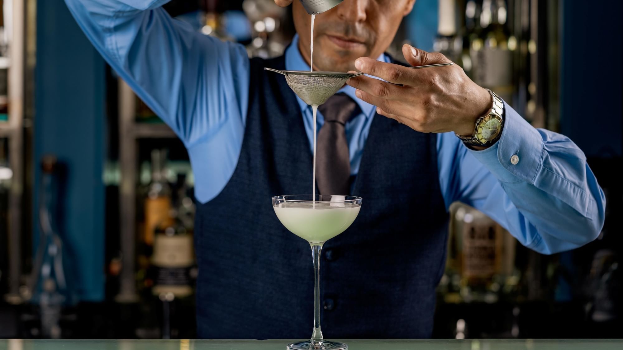 Barman in blue shirt and vest straining drink into a martini glass over a bar at Warwick Hotels and Resorts