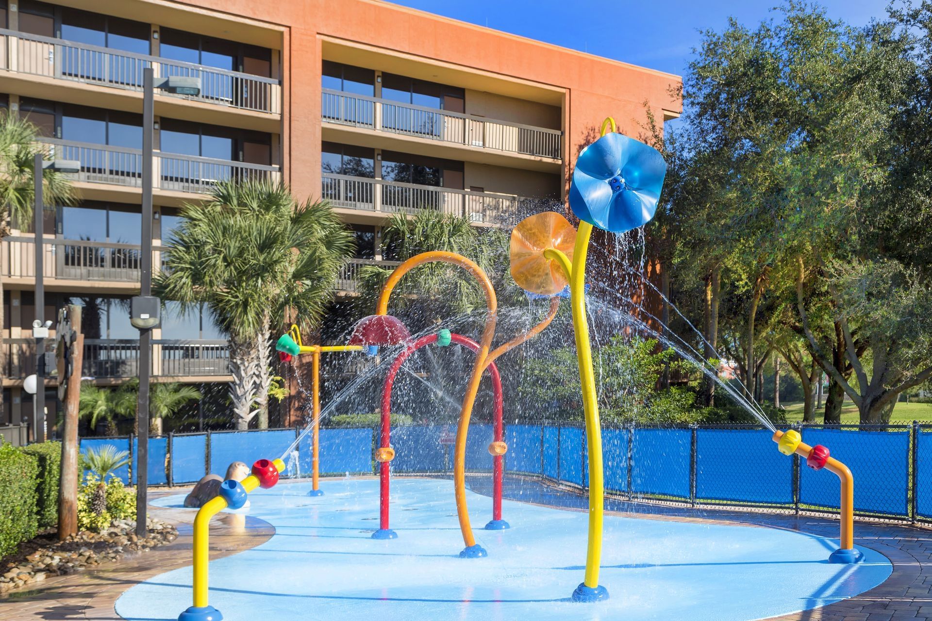 An interactive splash pad in front of a hotel building with colorful fountains spraying water surrounded by palm trees.