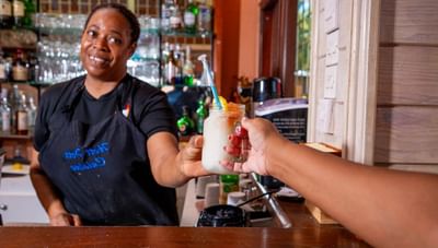 Smiling bartender in a black apron reaches out to hand a customer a frozen cocktail at Worthing Court Apartment