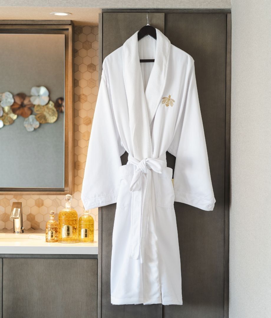 White robe with gold bee emblem hanging by a spa vanity, under amenity bottles and a floral mirror at Hotel X Toronto