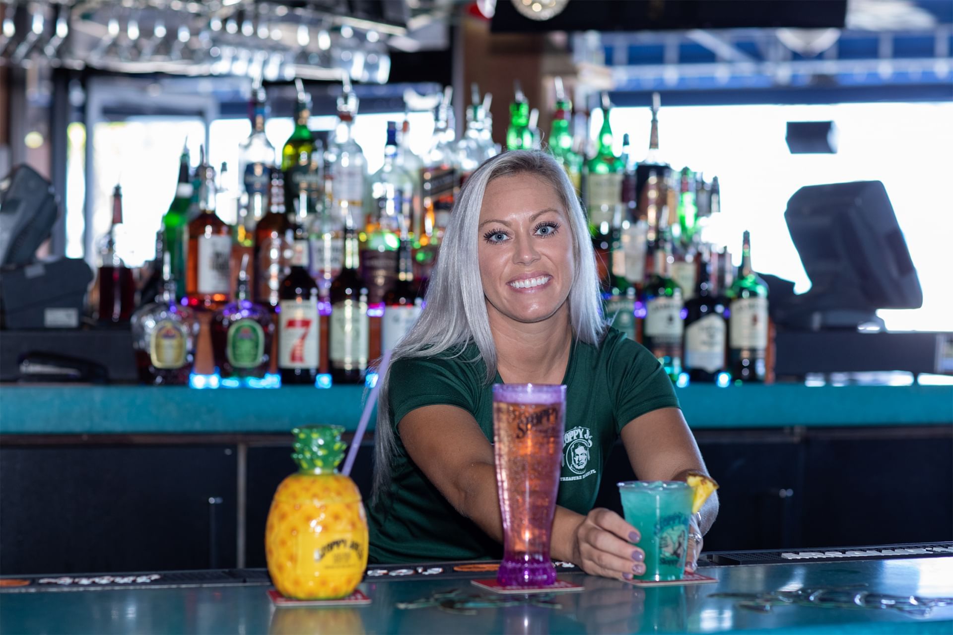 A smiling woman with long blonde hair behind a bar with various bottles and glasses, with three colorful drinks in front of her.