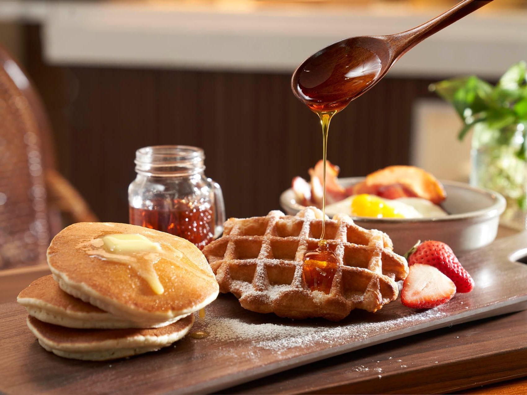 Breakfast buffet at Orchard Grand Court with waffles, pancakes, fruit, and a syrup jar on a rustic wooden platter