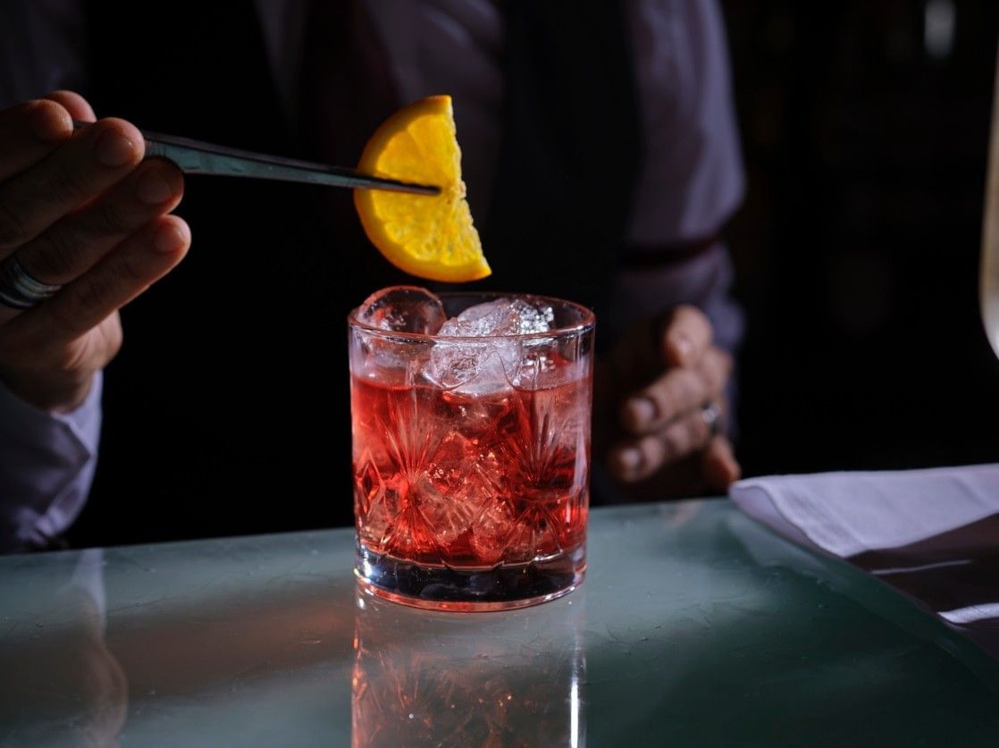 Bartender using tweezers by a bar to place an orange slice in a red cocktail at The Capital Hotel, Apartments and Townhouse