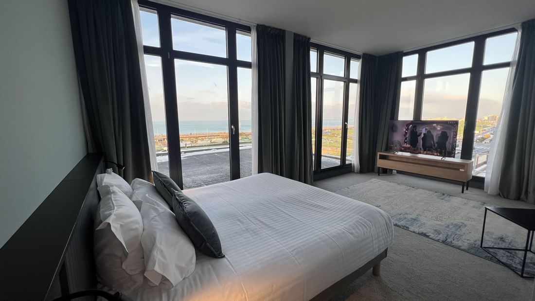 Chambre avec lit, télévision, et vue sur l'océan à travers de grandes fenêtres.