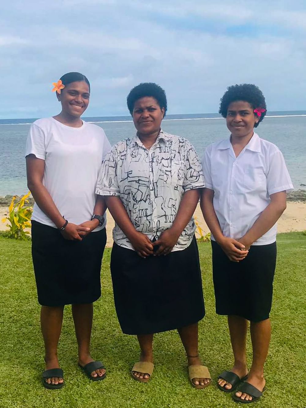 Three hospitality students in professional attire standing on beachfront lawn with ocean and palm trees in background.