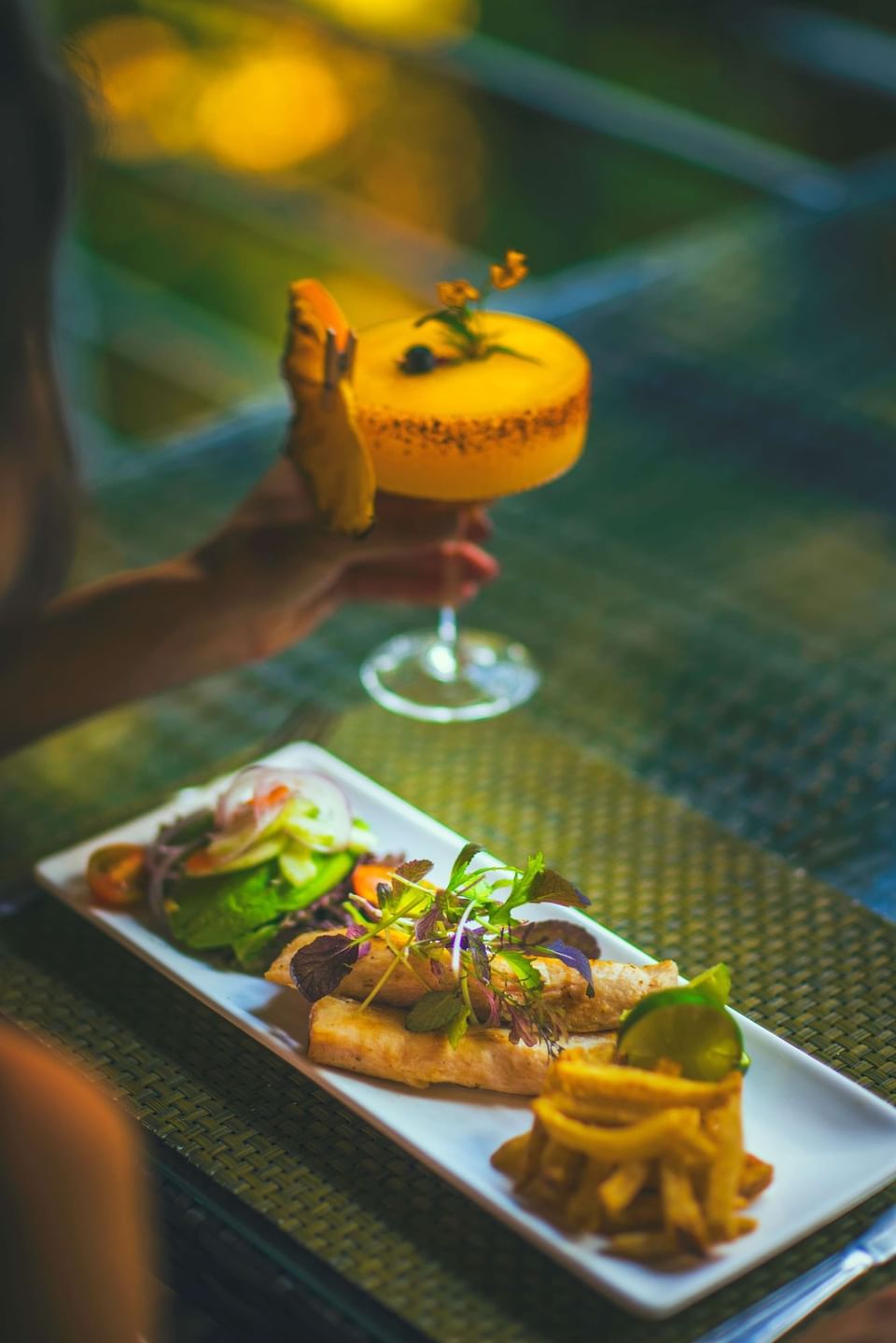 Yellow cocktail held above a grilled fish with salad and crisp fries in Sloth Restaurant at Jungle Vista Boutique Hotel