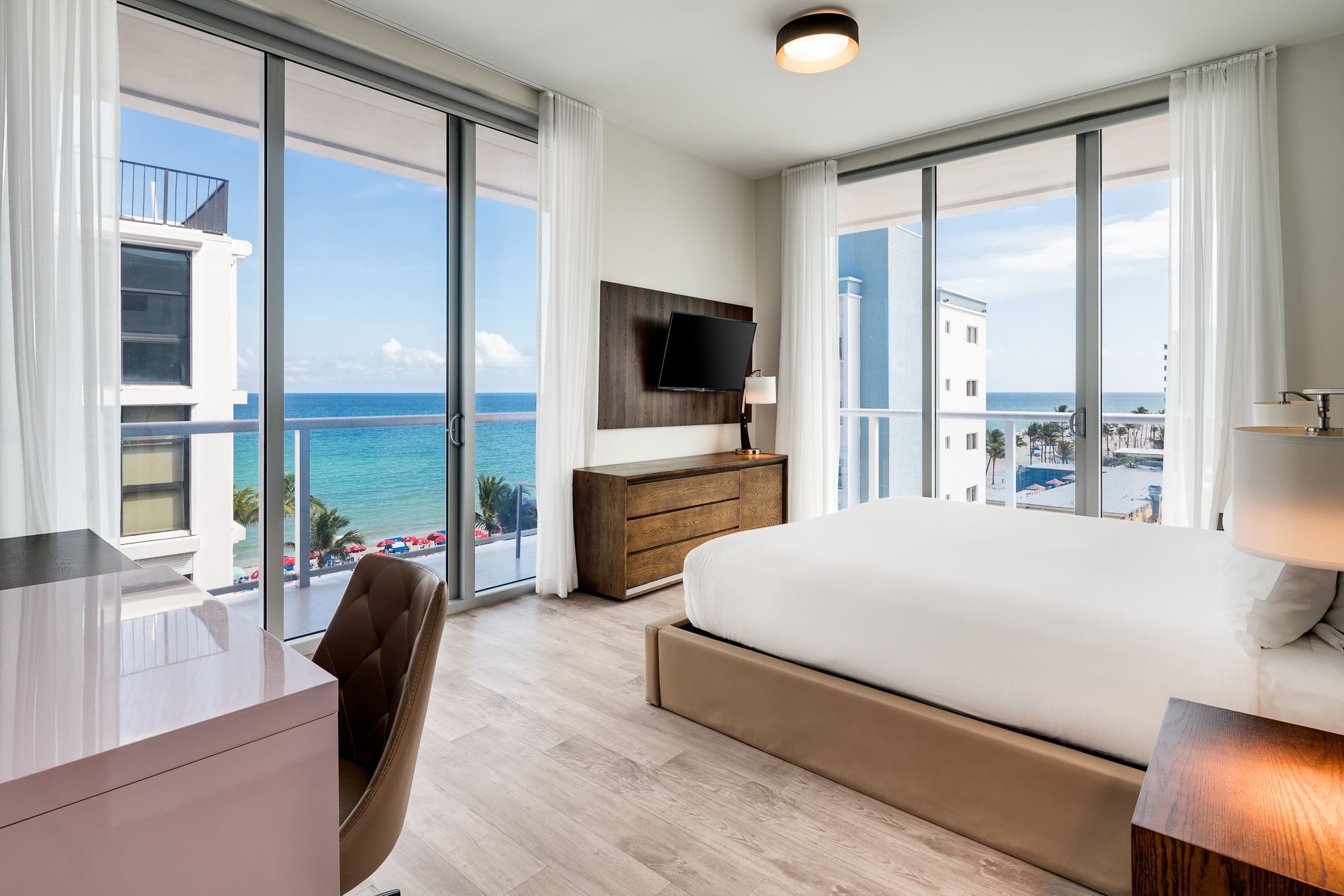 A wide-angle view of a hotel room with a bed, desk, TV, and ocean view.