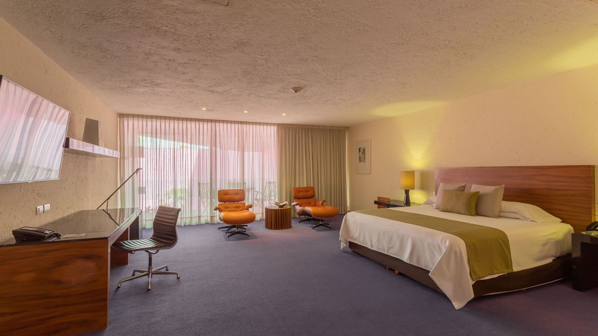 Habitación de hotel con cama grande, sillas naranjas, escritorio y televisor en la pared.
