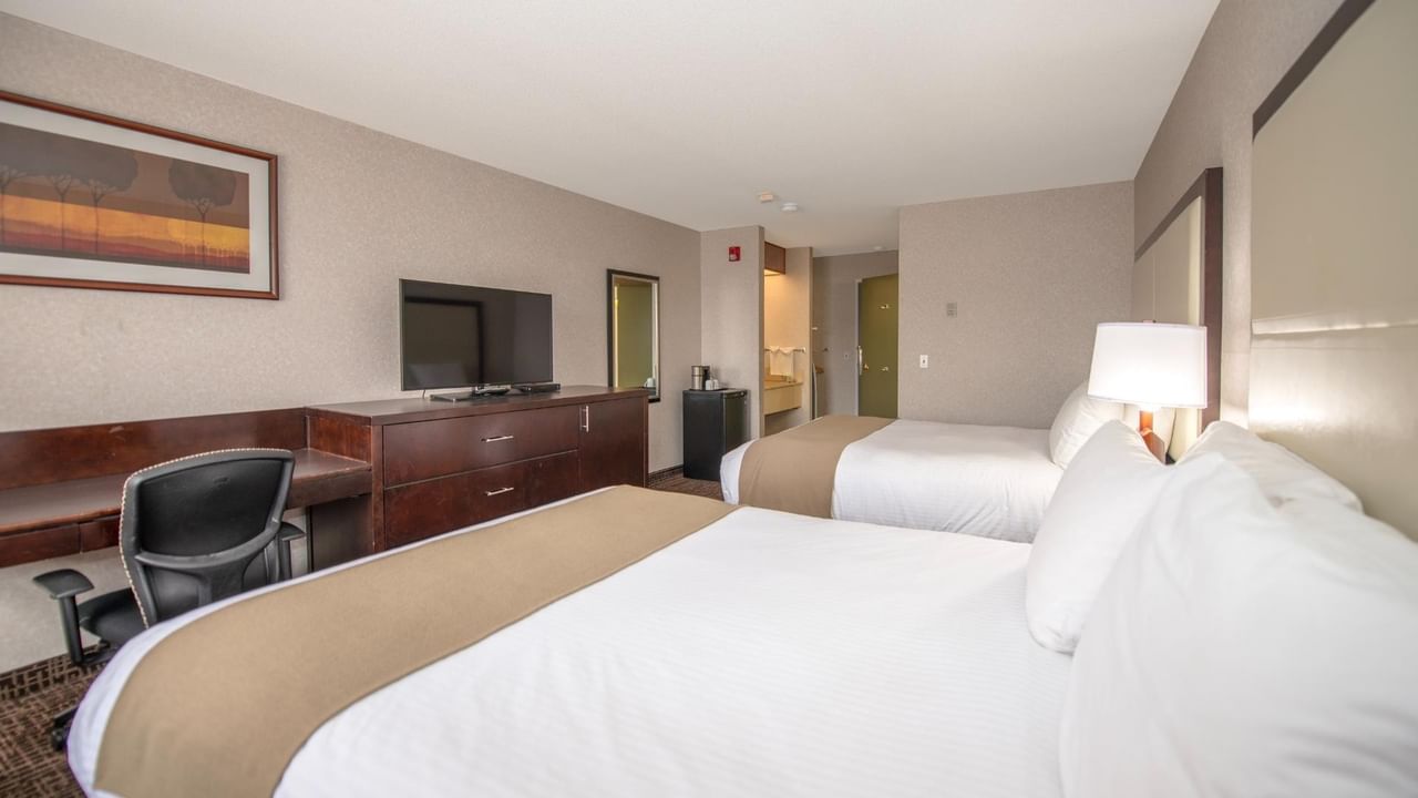 A hotel room with two beds, a desk, a chair, a TV, a lamp, and a picture frame.