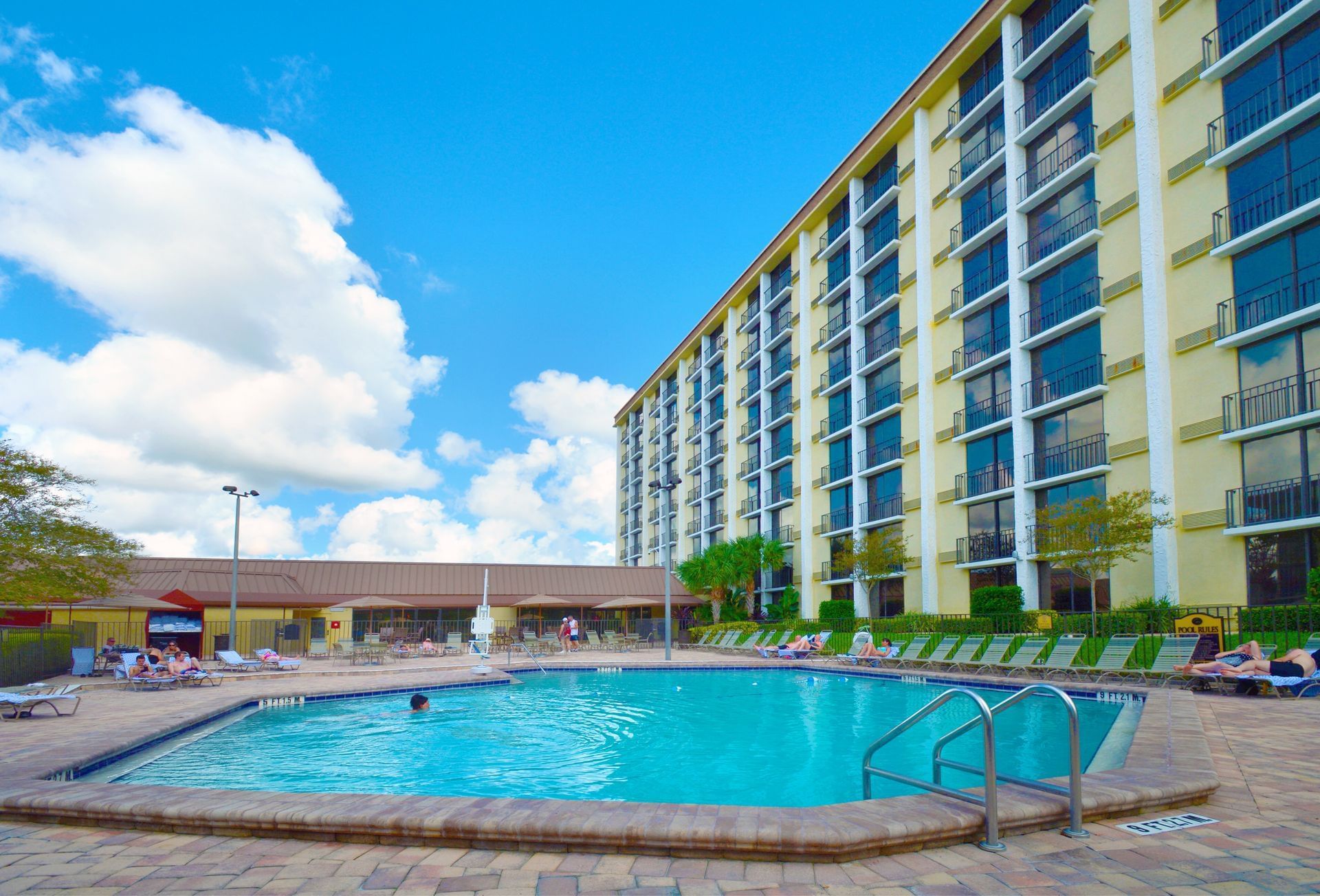 A large yellow hotel building beside a crystal clear pool surrounded by lounge chairs beneath a blue sky with clouds. 