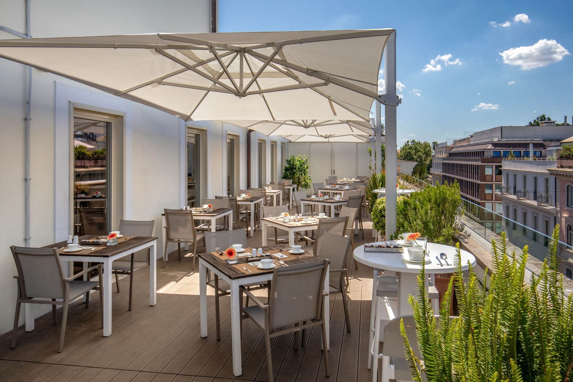 Outdoor café terrace with tables under large umbrellas, surrounded by greenery and a bright sky at The Glam Hotel