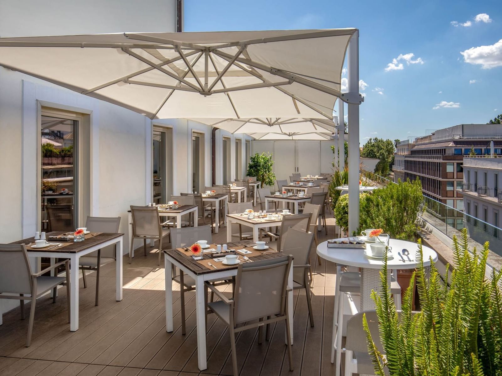 Outdoor café terrace with tables under large umbrellas, surrounded by greenery and a bright sky at The Glam Hotel