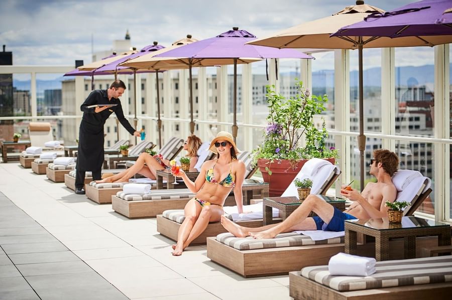 Guests on lounge chairs by a server under purple umbrellas near a glass rail at Warwick Denver