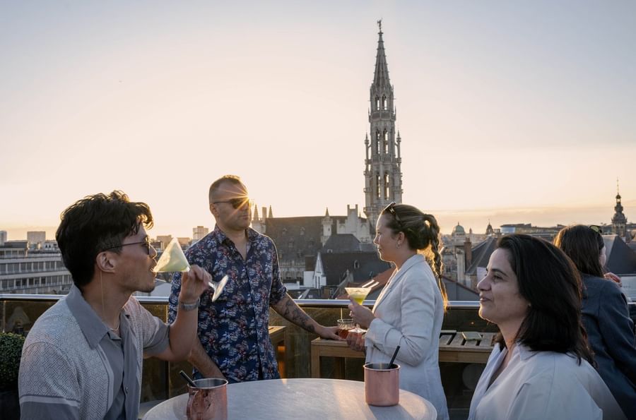 Vier gasten genieten van drankjes op een dakterras met uitzicht op de Brusselse skyline bij zonsondergang