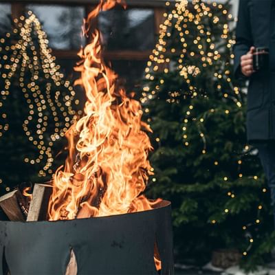 Person hält Getränk neben brennendem Feuerkorb vor leuchtendem Weihnachtsbaum.