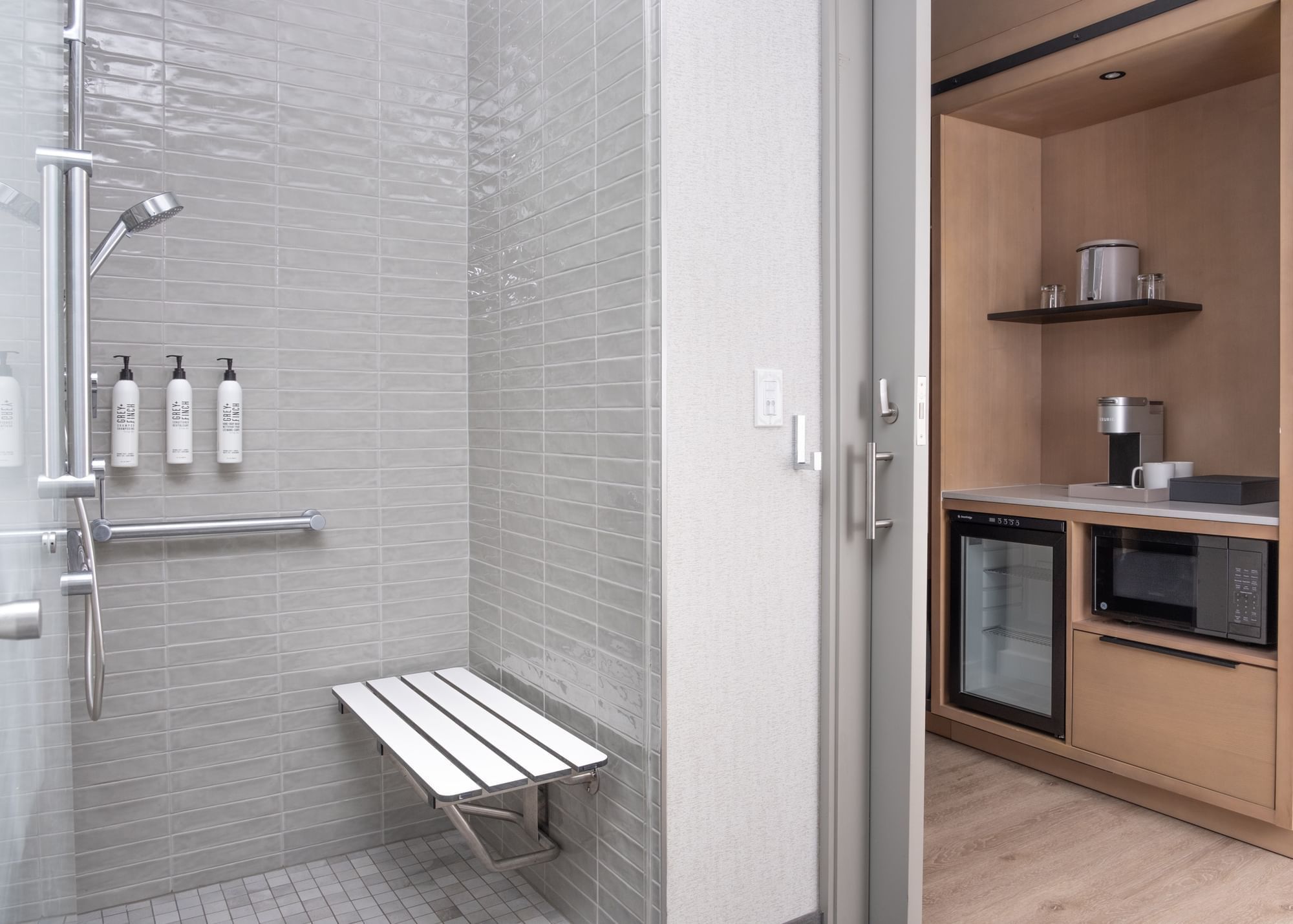 A modern bathroom with a walk-in shower, bench, and toiletries, adjacent to a small kitchenette with a mini fridge and microwave.