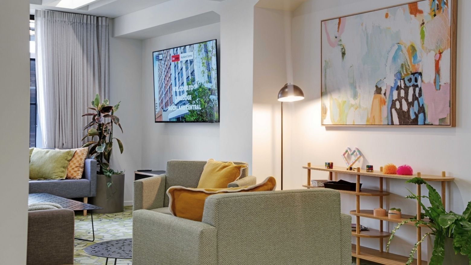Modern lounge area with gray sofas, yellow cushions, wall-mounted TV, and colorful artwork at UniLodge Auckland Central