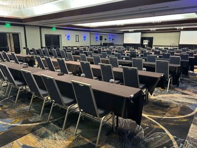 Conference room with rows of tables and blue chairs facing large screens in the Grand Slam Ballroom at Miami Lakes Hotel