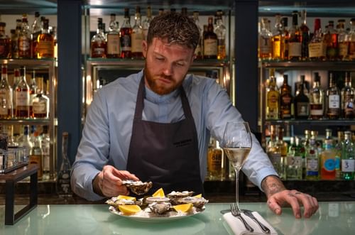 Bartender in a dark apron by shelves of liquor, preparing fresh oysters at The Capital Hotel, Apartments and Townhouse
