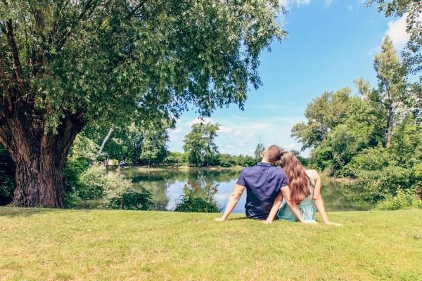 A couple sits on the bank of a river leaning on one another beneath a large tree. 