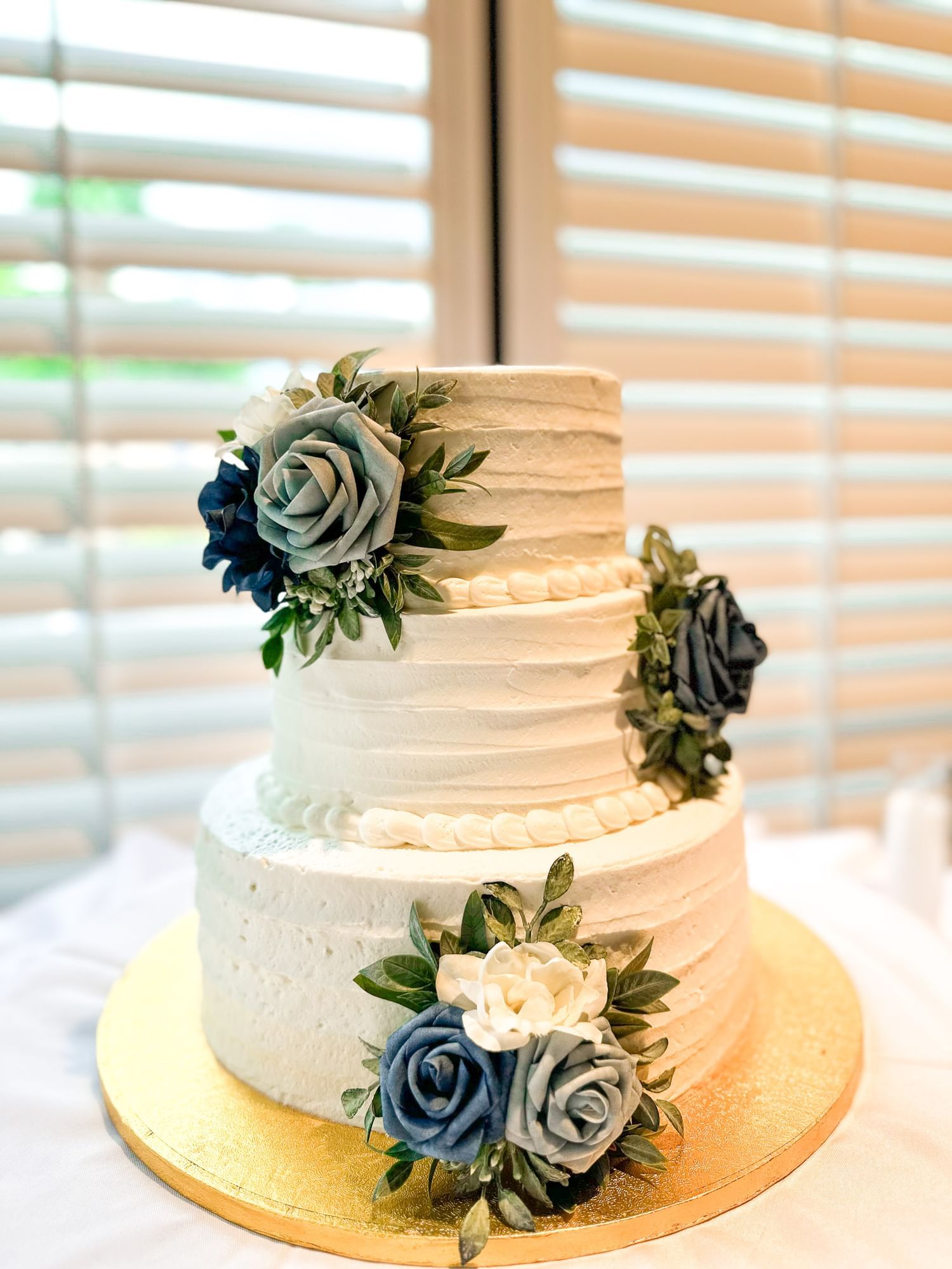 A three-tiered white wedding cake with blue and white flowers on a gold plate