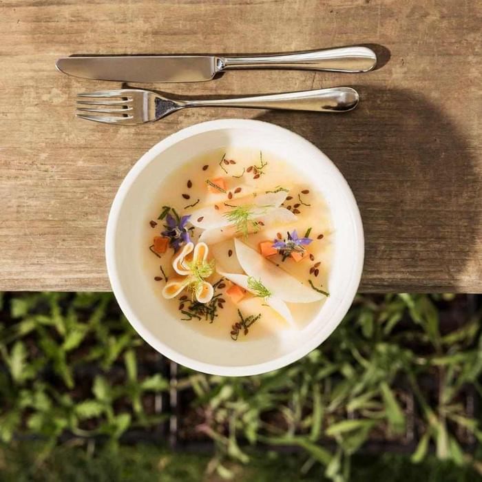 White bowl of soup with vegetables and herbs, knife and fork on wooden surface, greenery below.
