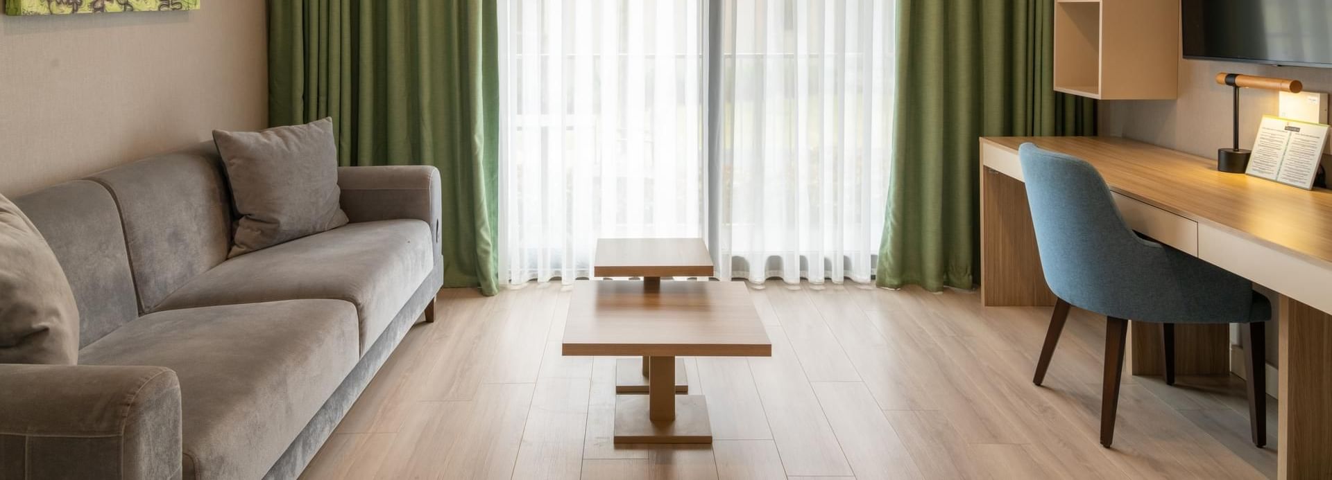 Modern guest suite at The Hub Hotels, with a grey sofa, light wood desk, and vibrant green window curtains