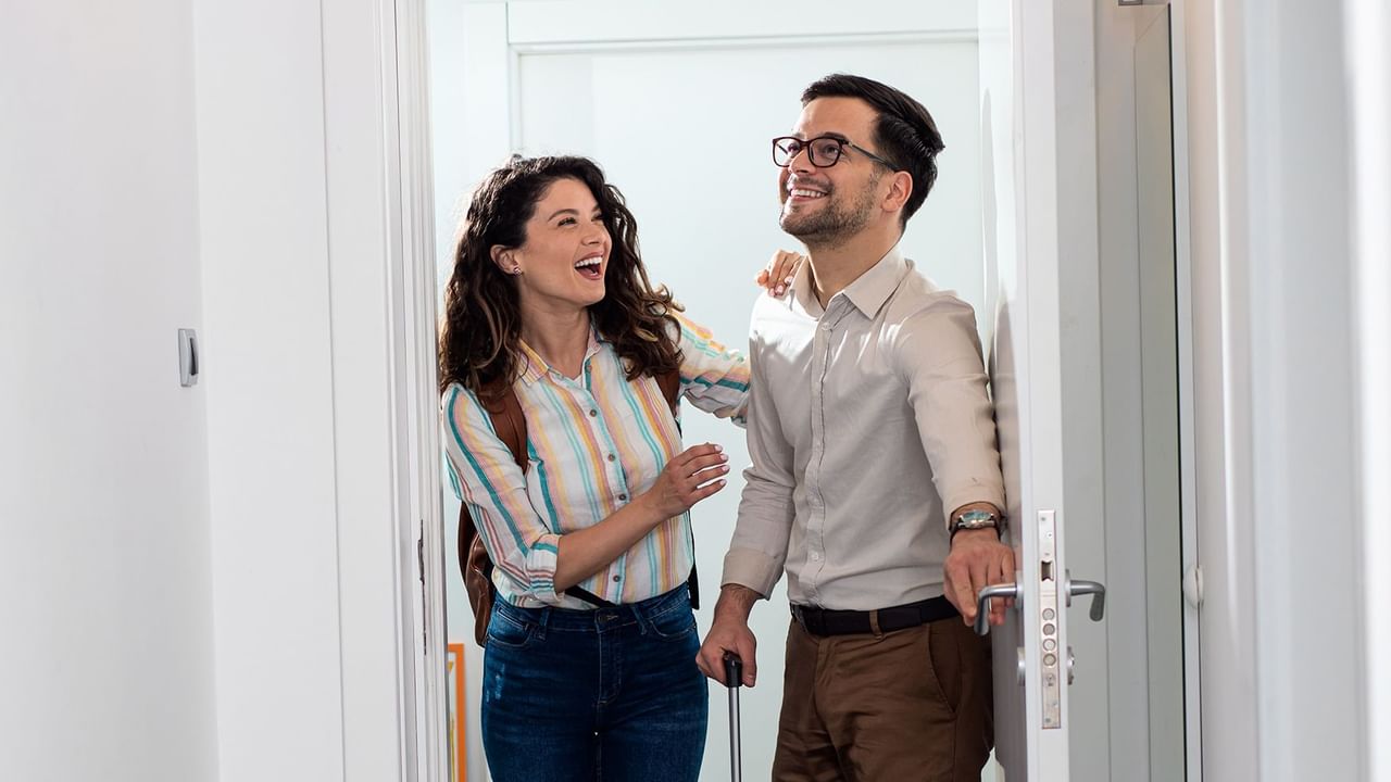 Couple entering hotel room, woman smiling, man holding the door.