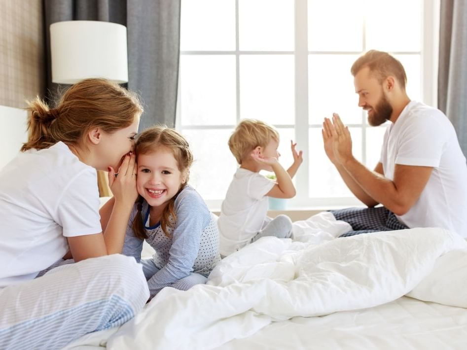 Family enjoying a playful morning at Blair Hotels, with a mother whispering to her smiling daughter on the bed