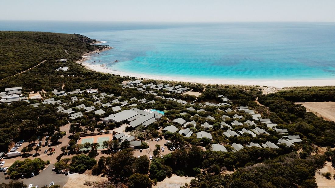 Aerial view of a coastal resort with white tents nestled among greenery beside a pristine turquoise beach and ocean.