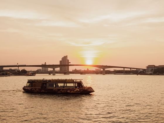 Scenic sunset over a river with a small boat in the foreground and a bridge near The Okura Prestige Bangkok