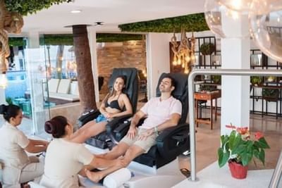 Two guests relaxing in massage chairs while receiving foot treatments in a bright spa lounge at Playa Los Arcos Puerto Vallarta