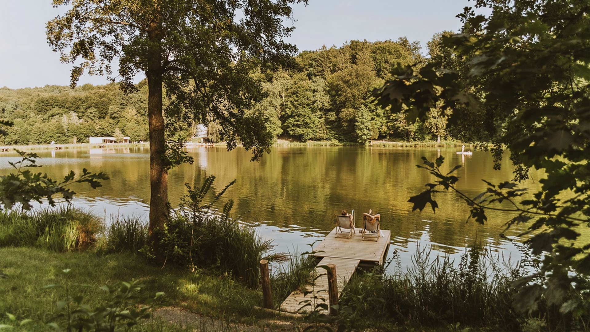 Zwei Personen sitzen auf Stühlen auf einer Holzplattform in einem See, umgeben von Bäumen.