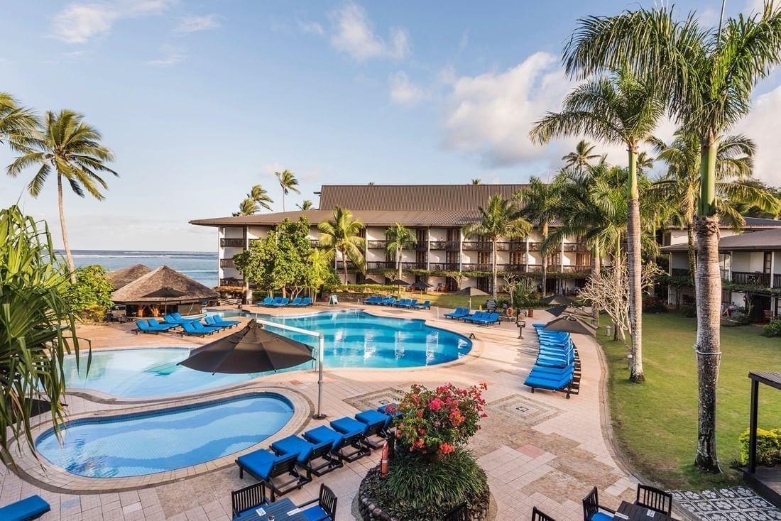 Blue lounge chairs by a large pool under tall palms near a multi-story wing at Warwick Fiji Resort and Spa