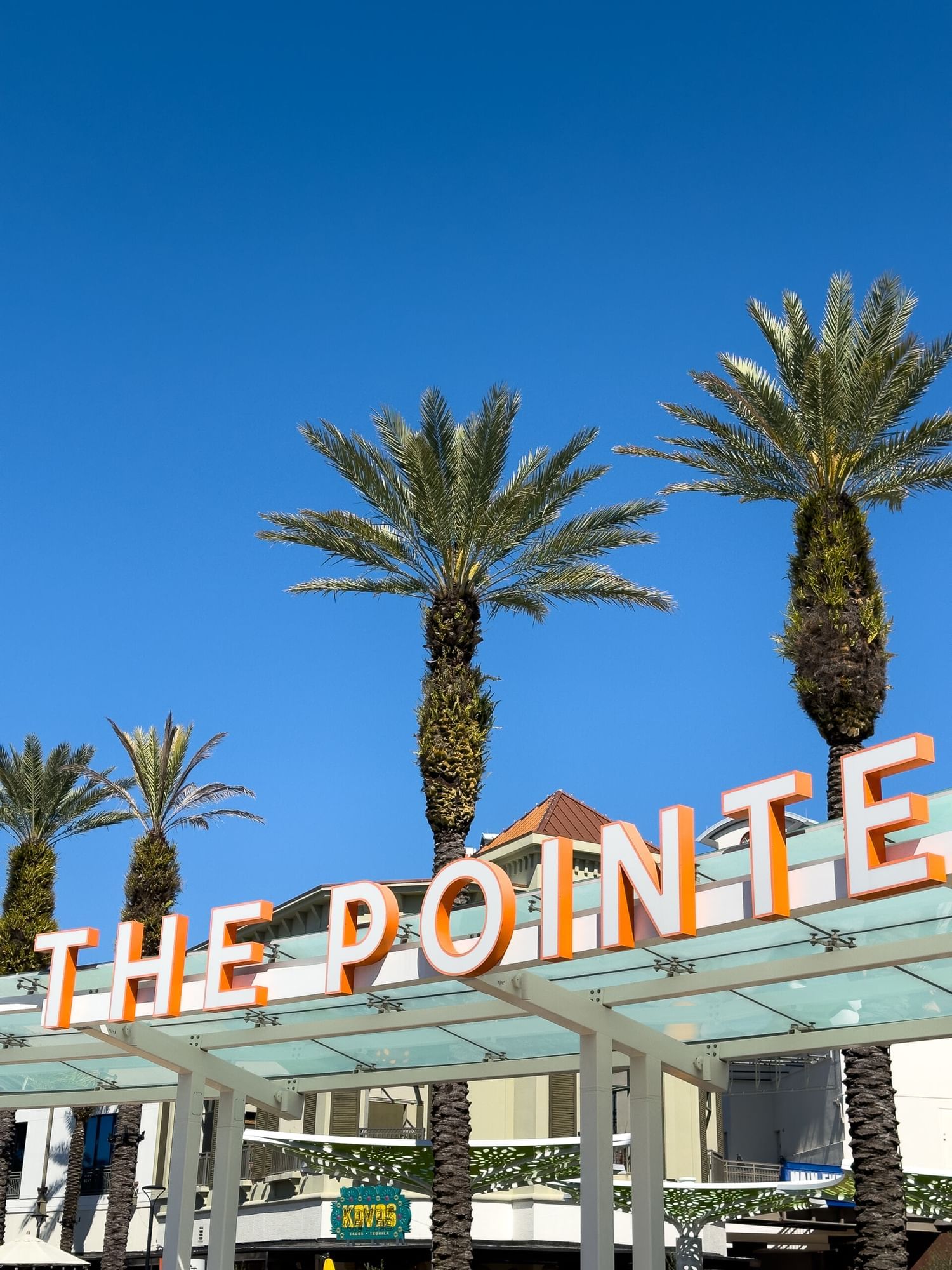 An awning with a sign that reads The Pointe in orange and white letters against a blue sky with palm trees and storefronts.