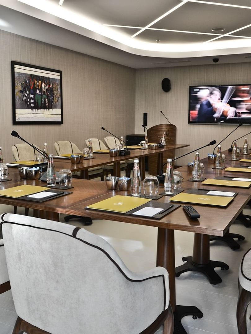Grey chairs by a wooden table surrounding a large TV screen under bright lights in Kugu Hall at Warwick Ankara