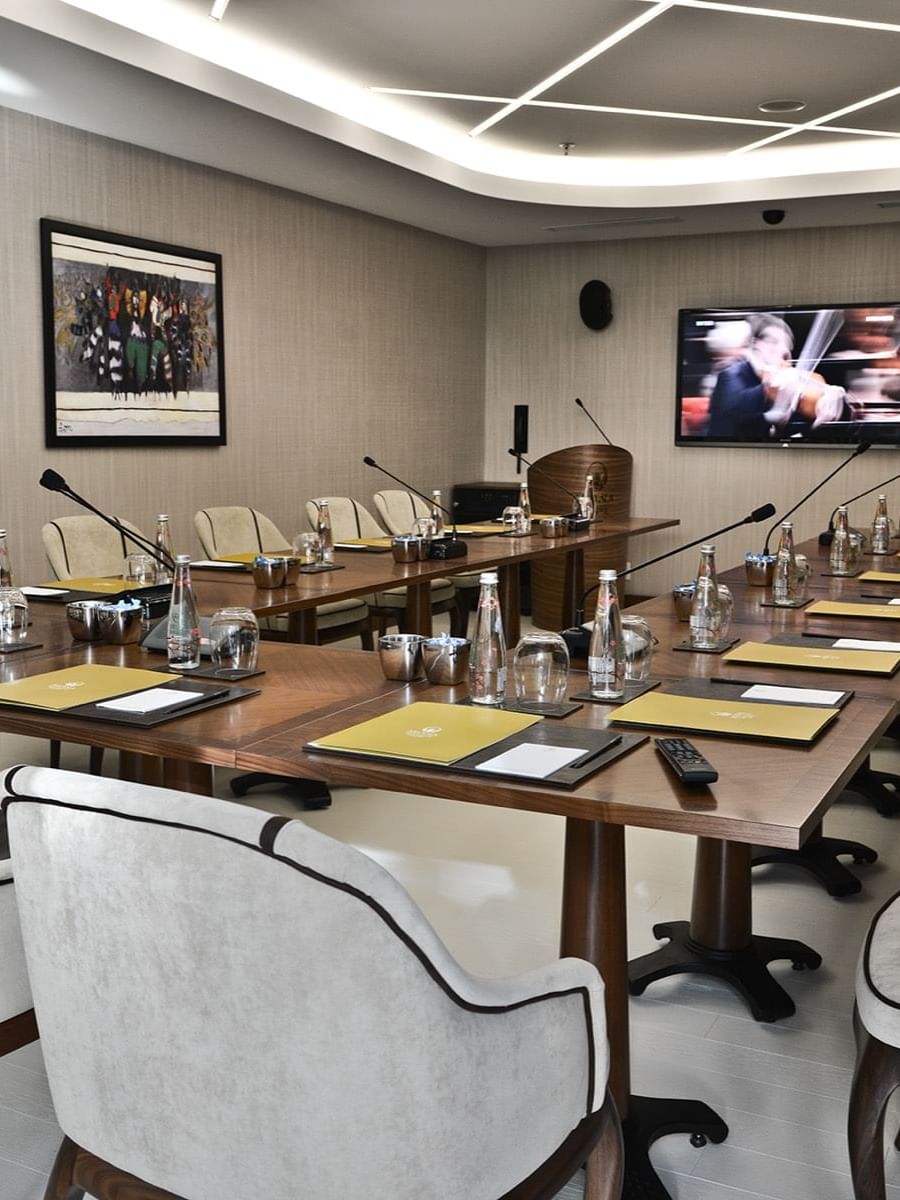 Grey chairs by a wooden table surrounding a large TV screen under bright lights in Kugu Hall at Warwick Ankara