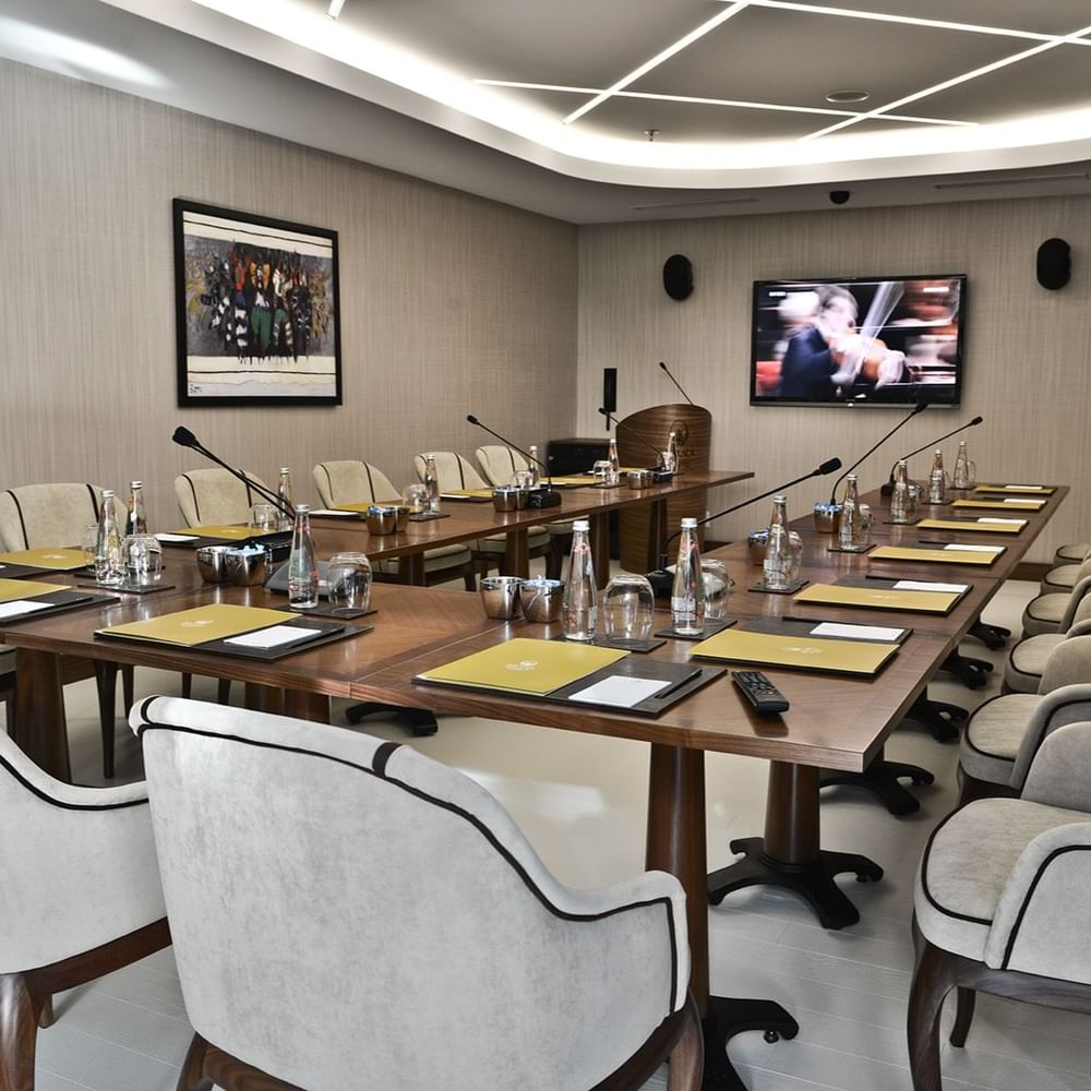 Grey chairs by a wooden table surrounding a large TV screen under bright lights in Kugu Hall at Warwick Ankara
