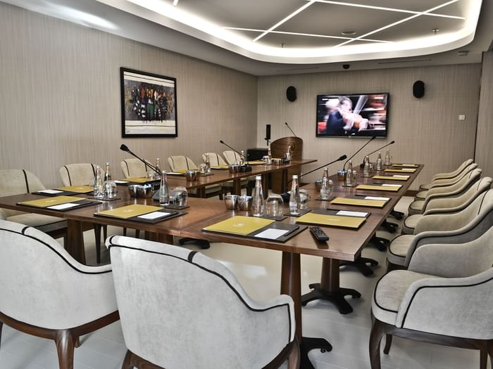 Grey chairs by a wooden table surrounding a large TV screen under bright lights in Kugu Hall at Warwick Ankara