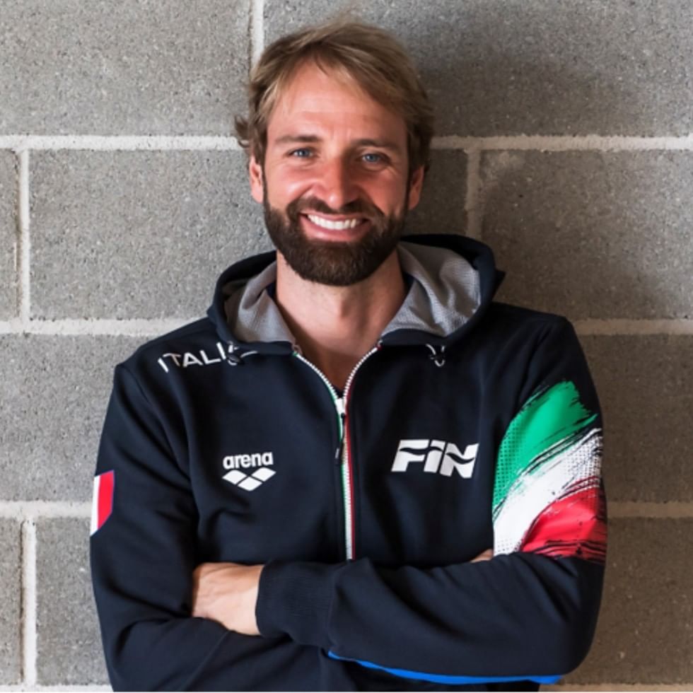 Man smiling with arms crossed wearing a blue hoodie with FINA logo and Italian flag colors.
