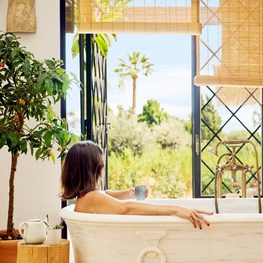 Mujer en bañera de mármol con vista a un jardín y palmeras, disfrutando de una taza.