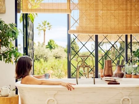Woman relaxing in a bathtub with a cup, surrounded by plants and natural light from large windows.