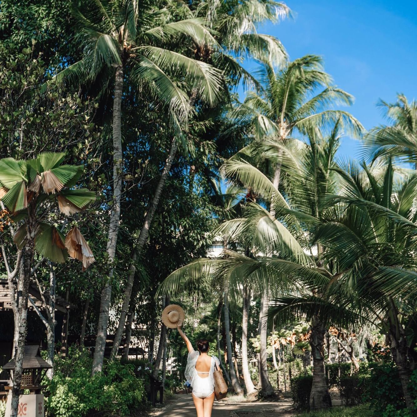 A woman wearing white costume is walking and enjoying the green pathway surrounded by trees in the resort