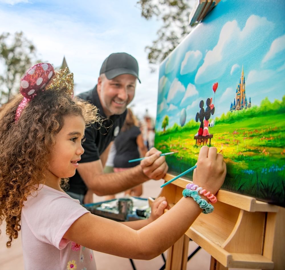 A man and a girl in Minnie ears paint at an easel with a painting of Mickey and Minnie sitting on a bench. 