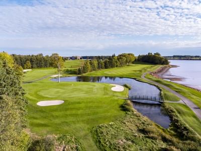Scenic golf green by a winding pond under a bright, cloudy sky, surrounded by trails near Rodd Brudenell River Resort