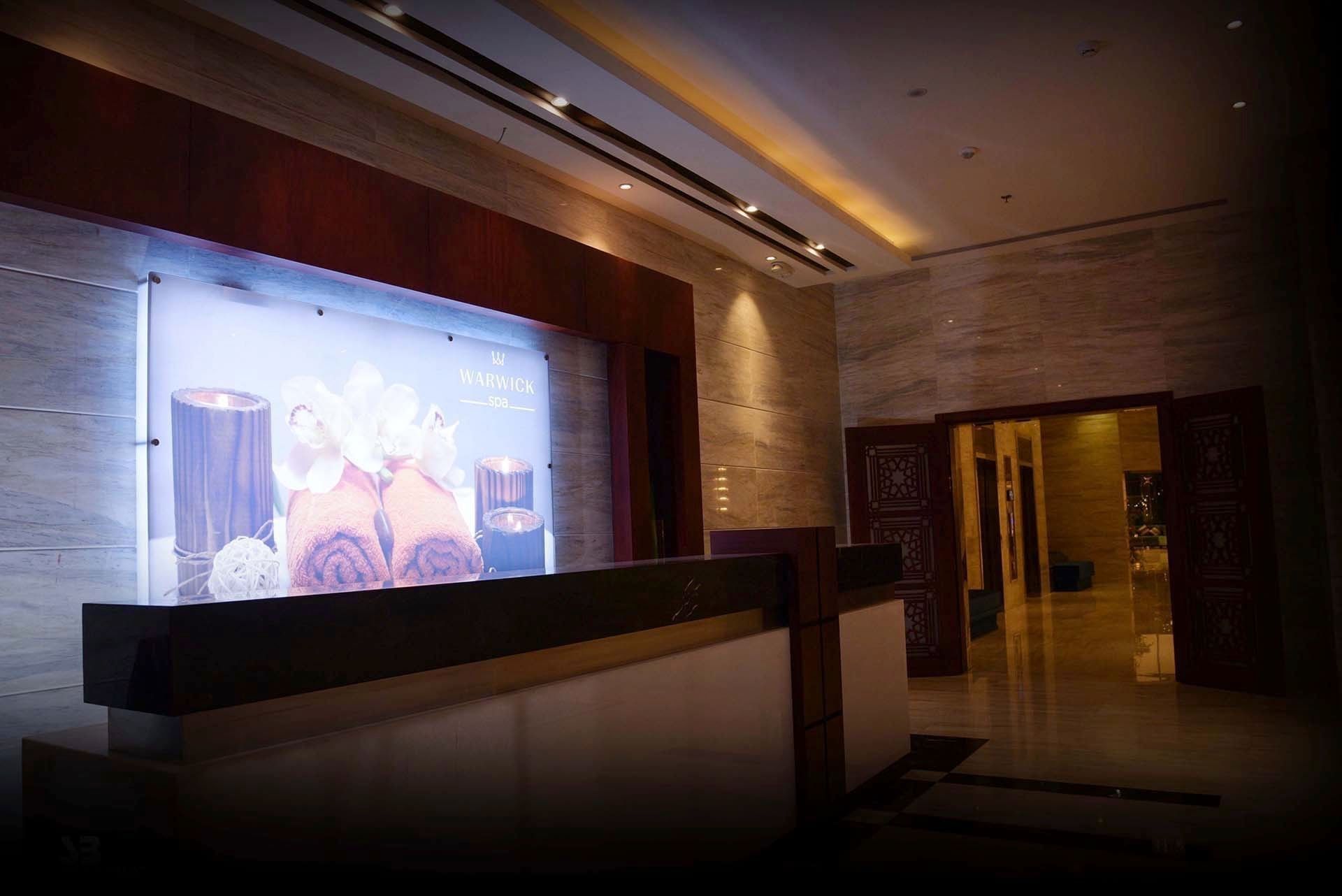 Dark wood spa reception desk under recessed lighting with a large orchid photo near a doorway at Warwick Al Khobar