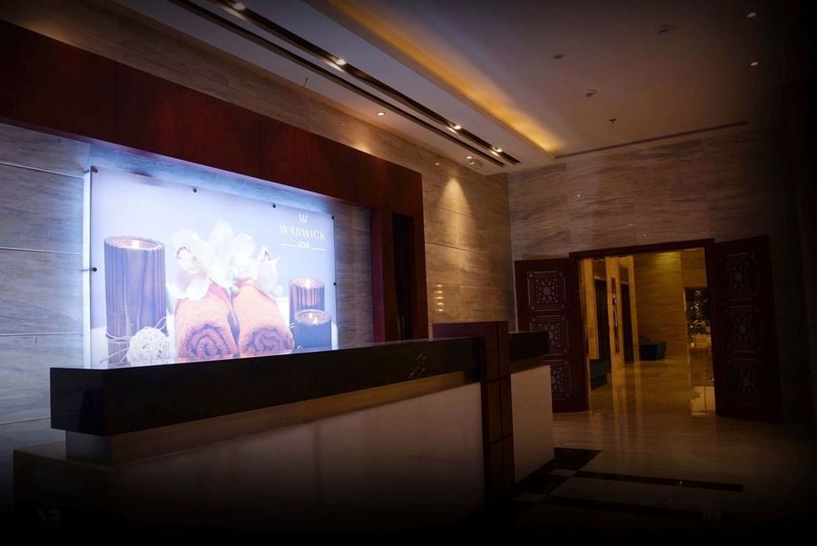 Dark wood spa reception desk under recessed lighting with a large orchid photo near a doorway at Warwick Al Khobar