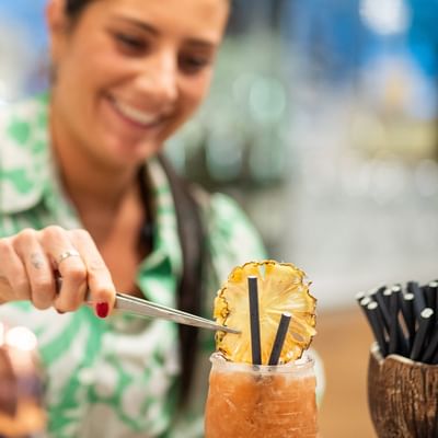 Frau mit grünem Shirt mischt einen Cocktail mit Ananasstück und Strohhalm.