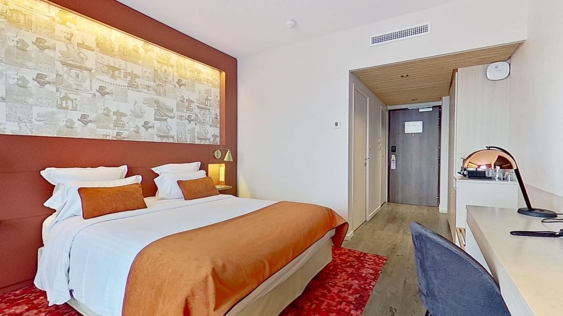 A modern hotel room with a large bed, a desk, and a chair near a hallway.