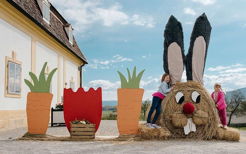 Kinder spielen neben einer großen Osterhasen-Figur aus Heu mit bunter Osterdekoration im Schlosspark.